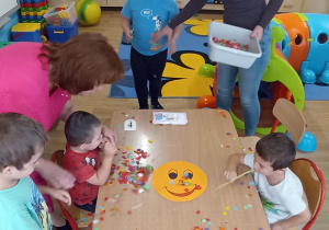 Dzieci z grupy 4 rozdmuchują przez słomki bibułę z emotki przedstawiającej radość.